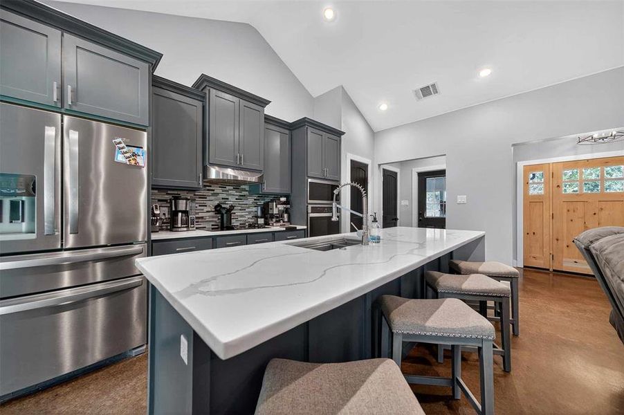 Kitchen with stainless steel appliances, an island with sink, backsplash, light stone counters, and gray cabinets Kitchen with stainless steel appliances, an island with sink, backsplash, light stone counters, and gray cabinets