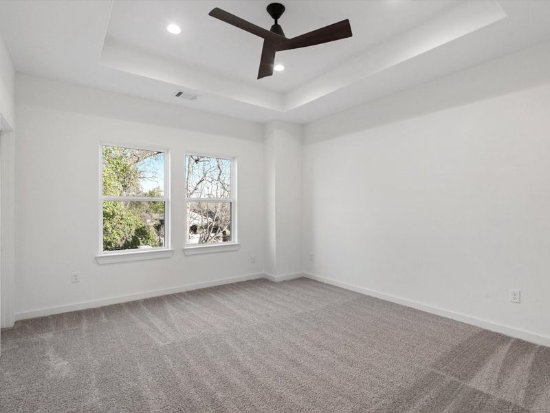 This primary bedroom features a spacious area with plush carpeting, large windows for natural light, and a modern ceiling fan. The neutral color palette and tray ceiling add a touch of elegance, perfect for a bedroom or office.