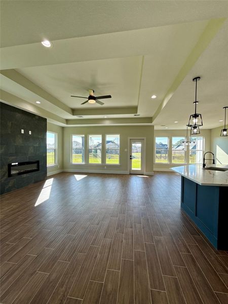 This spacious living area features wood-look tile flooring, a modern ceiling fan, and abundant natural light from large windows. It includes an elegant fireplace and an open kitchen with a sleek island, perfect for entertaining. This spacious living area features wood-look tile flooring, a modern ceiling fan, and abundant natural light from large windows. It includes an elegant fireplace and an open kitchen with a sleek island, perfect for entertaining.