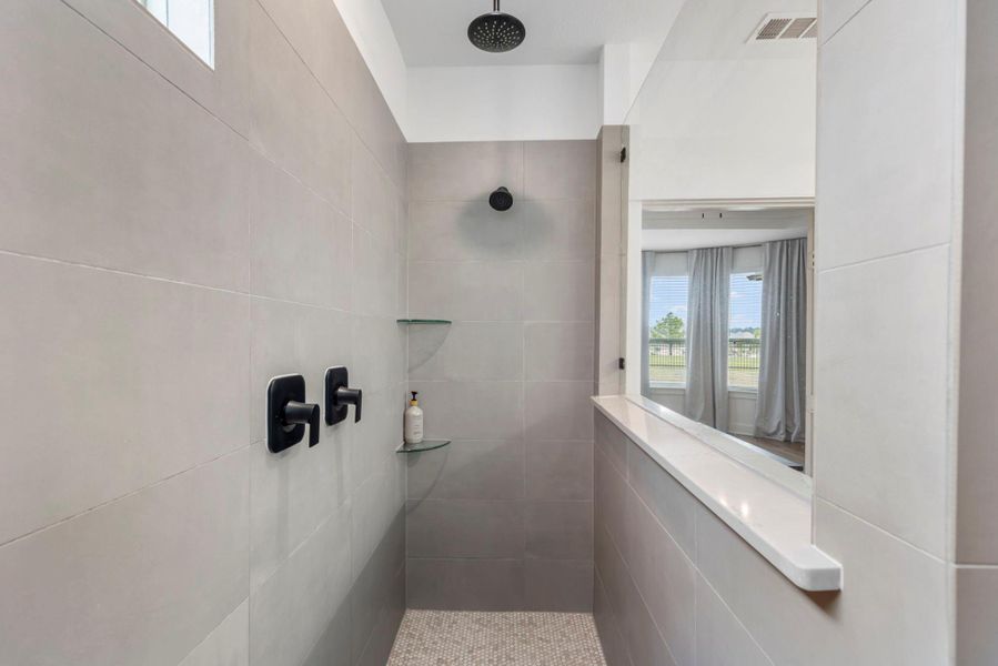 This photo showcases a modern shower with sleek gray tiles, a rainfall showerhead, and matte black fixtures. It features built-in glass shelves and an open view to a room with large windows, providing natural light and a spacious feel.