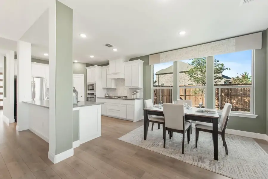 Open-plan dining area and kitchen with white cabinets, hardwood floors, and large windows overlooking a backyard.