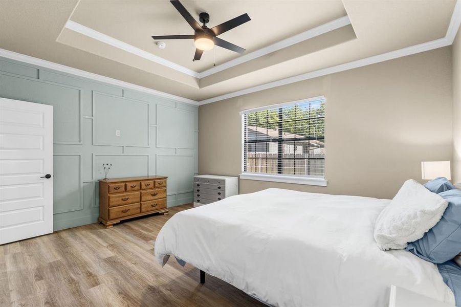 Room featuring a tray ceiling with a ceiling fan, wood-finish flooring, a paneled accent wall, a multi-paneled door with dark hardware, and a window with blinds