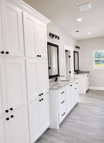 Bright bathroom featuring extensive white cabinetry with black hardware, dual vanity sinks with light-toned countertops, and wood-finish flooring Bright bathroom featuring extensive white cabinetry with black hardware, dual vanity sinks with light-toned countertops, and wood-finish flooring