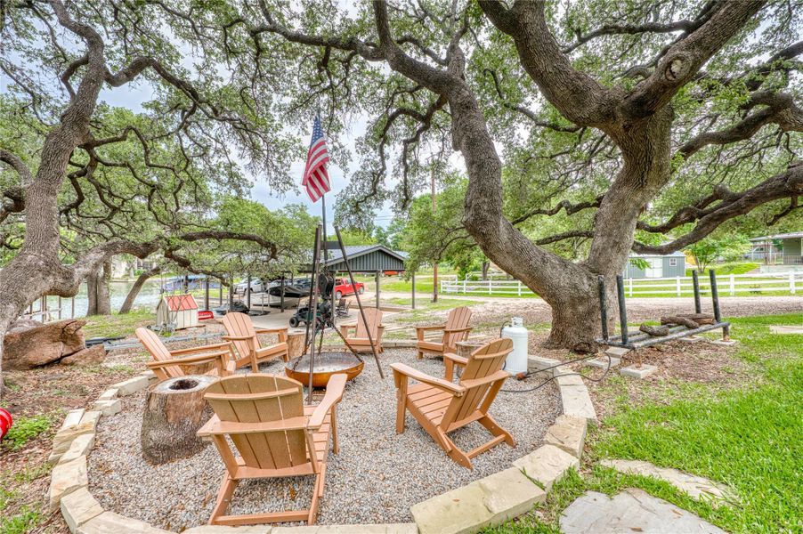 Outdoor fire pit area featuring a gravel base, stone border, and mature trees providing natural shade