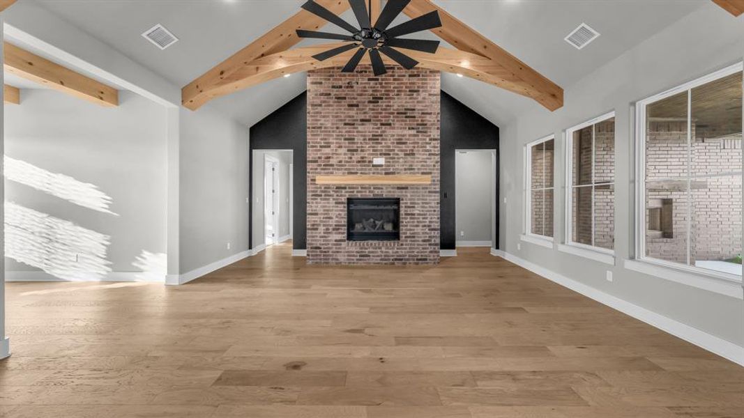 Spacious living area featuring a vaulted ceiling with exposed wood beams, a brick fireplace with a wood mantel, wood-finish flooring, and multiple large windows Spacious living area featuring a vaulted ceiling with exposed wood beams, a brick fireplace with a wood mantel, wood-finish flooring, and multiple large windows
