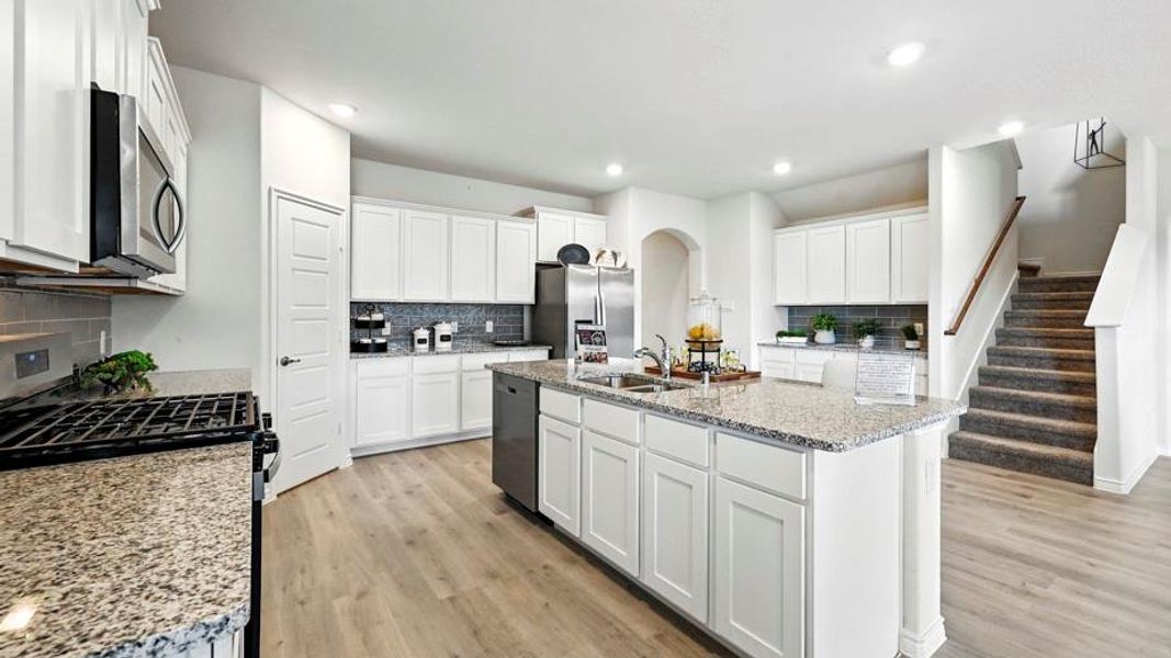 This kitchen features light-colored countertops, white cabinetry, a kitchen island with a double sink, stainless steel appliances, and wood-look flooring