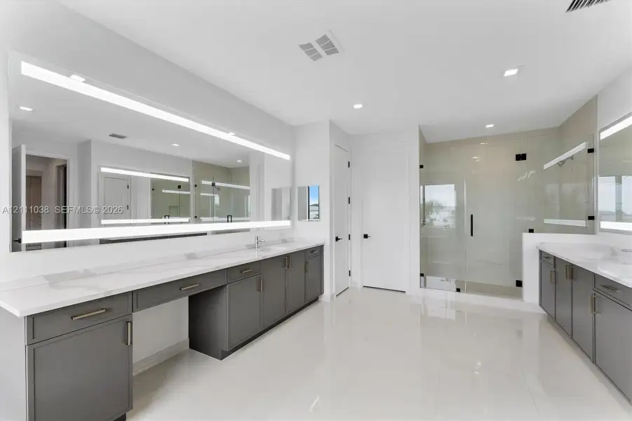 Dual vanities featuring charcoal cabinetry and white stone countertops, expansive wall-to-wall LED-backlit mirrors, polished tile flooring, and a glass-enclosed shower with tile surround