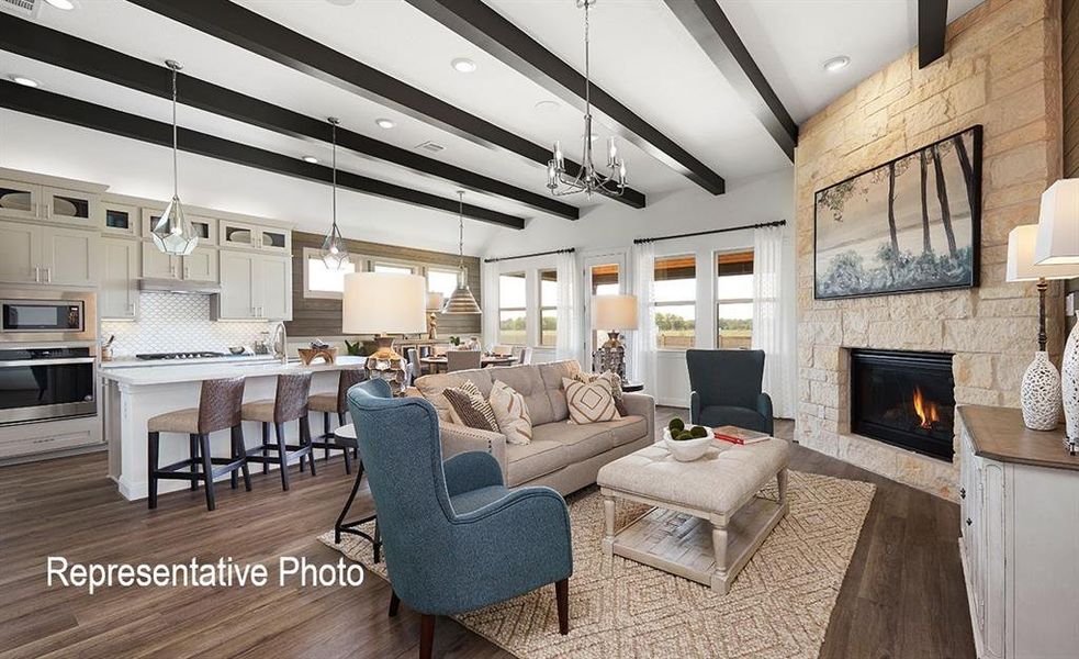 Open-concept living space featuring wood-finish flooring, exposed ceiling beams, a stone-faced fireplace, and an integrated kitchen with an island and built-in appliances