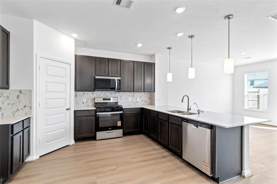 This spacious kitchen boasts high ceilings, Woodmont Dakota cabinets in a Smoke stain, Silestone Miami White countertops, stainless steel Whirlpool appliances, a modern Daltile backsplash, recessed lighting, pendant lighting, an extended breakfast bar countertop, and a pantry—all overlooking the expansive Great Room.