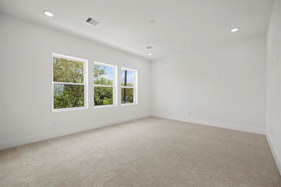 This bright and spacious Primary Bedroom is located on the 3rd level.  It has neutral carpeting and white walls. It features three large windows bringing in natural light and has recessed lighting in the ceiling.