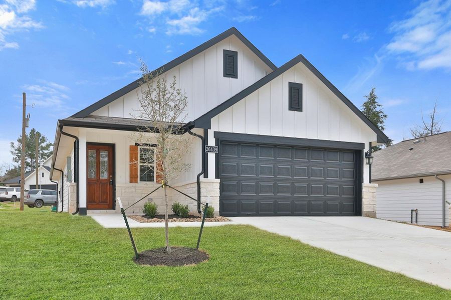 The clean, white exterior contrasts with   black trim. It includes a two-car garage, a welcoming front porch with a wooden door, and a neatly landscaped yard with a newly planted tree. Ideal for contemporary living.