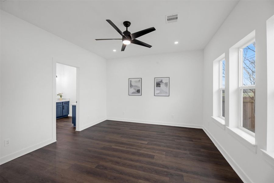Bright, spacious room with dark wood flooring, modern ceiling fan, and large windows for natural light. Adjacent to a room with blue cabinetry, ideal for versatile use.
