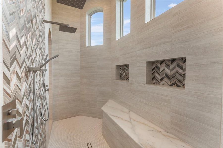 This is a spacious, modern walk-in shower is a dream shower. Featuring herringbone accent tiling, dual showerheads, built-in shelving, and tall windows for natural light.