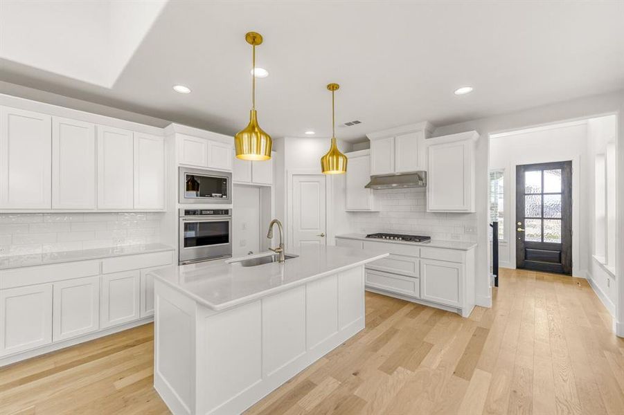 Kitchen featuring white cabinets, light wood-type flooring, stainless steel appliances, pendant lighting, and a kitchen island with sink