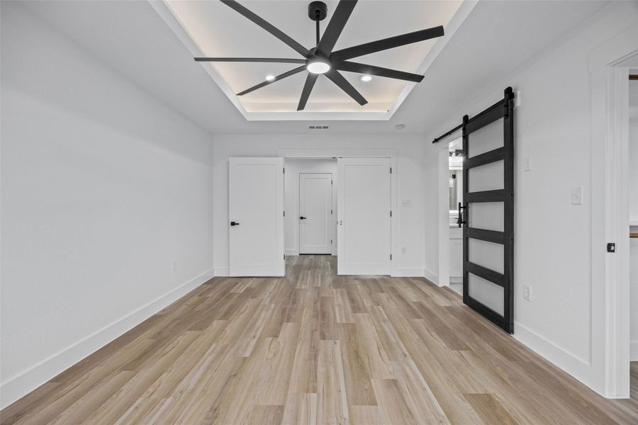 Spacious primary bedroom featuring wood-finish flooring, a contemporary ceiling fan, and a tray ceiling with recessed lighting Spacious primary bedroom featuring wood-finish flooring, a contemporary ceiling fan, and a tray ceiling with recessed lighting