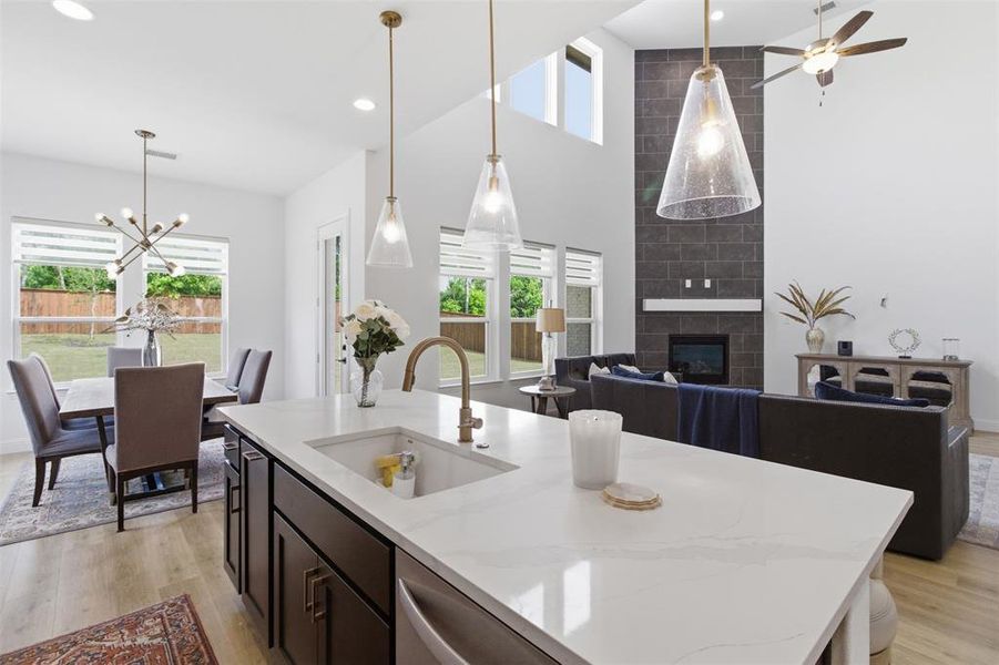 Open-concept living space with a kitchen island featuring a white countertop and undermount sink, pendant lighting, a two-story fireplace with dark tile surround, and wood-finish flooring