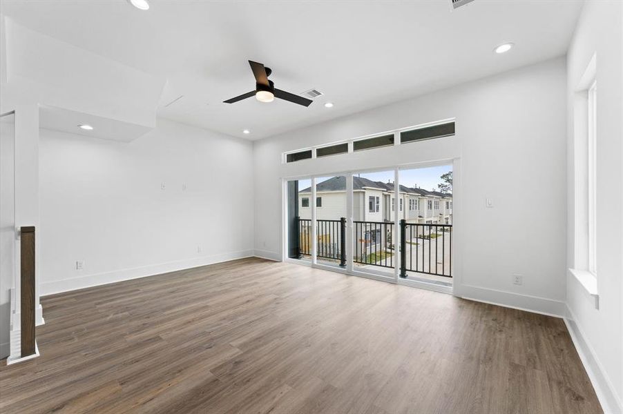 This room features a modern, open design with large windows and a sliding glass door leading to a balcony, providing ample natural light. The space is accented by recessed lighting, a ceiling fan, and sleek wood flooring, creating a bright and inviting atmosphere.