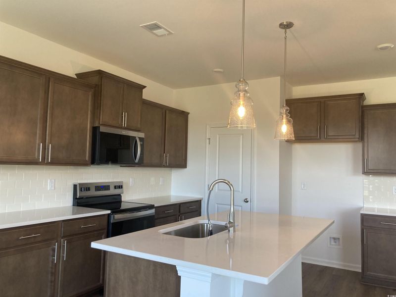 Kitchen with decorative backsplash, appliances with stainless steel finishes, dark brown cabinets, hanging light fixtures, and an island with sink