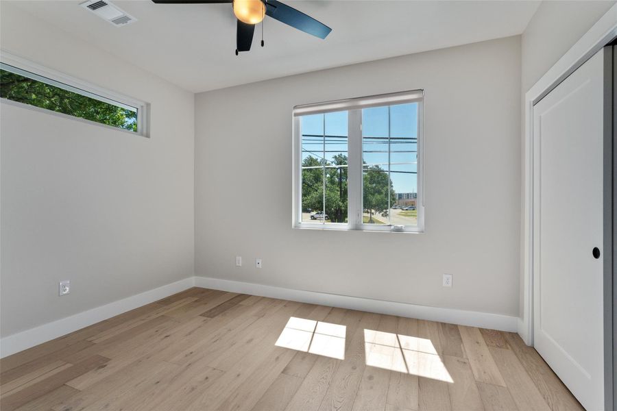 Light and airy secondary bedroom featuring wood-finish flooring, a large window with white framing, a high horizontal window, and a ceiling fan with integrated lighting. This secondary bedroom features a full bathroom. Light and airy secondary bedroom featuring wood-finish flooring, a large window with white framing, a high horizontal window, and a ceiling fan with integrated lighting. This secondary bedroom features a full bathroom.