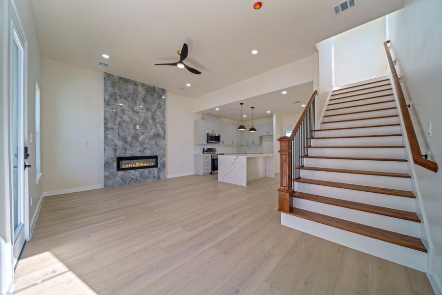 This modern open-plan living space features light LVP, a sleek fireplace, and a stylish ceiling fan. The kitchen boasts a large island with elegant pendant lighting. A staircase with wooden handrails leads to the upper floor, enhancing the home's bright and airy feel. This modern open-plan living space features light LVP, a sleek fireplace, and a stylish ceiling fan. The kitchen boasts a large island with elegant pendant lighting. A staircase with wooden handrails leads to the upper floor, enhancing the home's bright and airy feel.