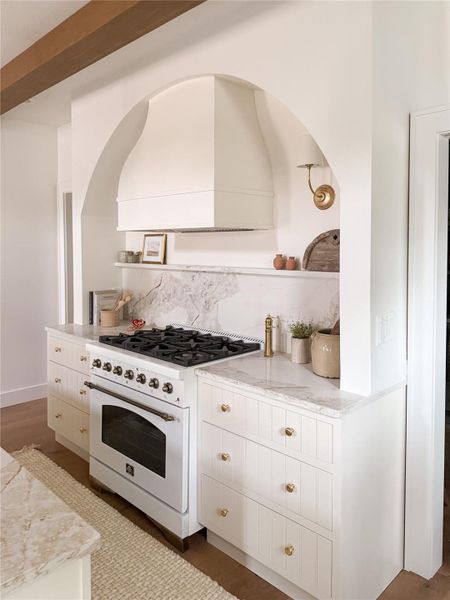 Kitchen featuring an arched detailed wall, custom range hood, a large Forno white with antique brass 6 burner gas range, white cabinetry with brass hardware, high-end quartz countertops with a matching backsplash, and white oak flooring Kitchen featuring an arched detailed wall, custom range hood, a large Forno white with antique brass 6 burner gas range, white cabinetry with brass hardware, high-end quartz countertops with a matching backsplash, and white oak flooring