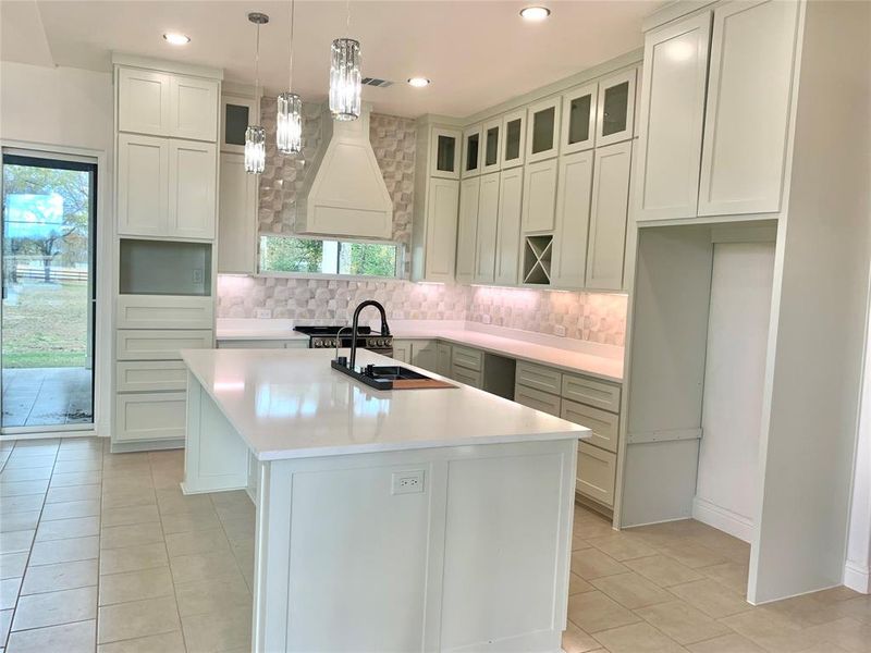 Kitchen with a kitchen island with sink, decorative light fixtures, backsplash, light tile patterned flooring, and glass insert cabinets Kitchen with a kitchen island with sink, decorative light fixtures, backsplash, light tile patterned flooring, and glass insert cabinets