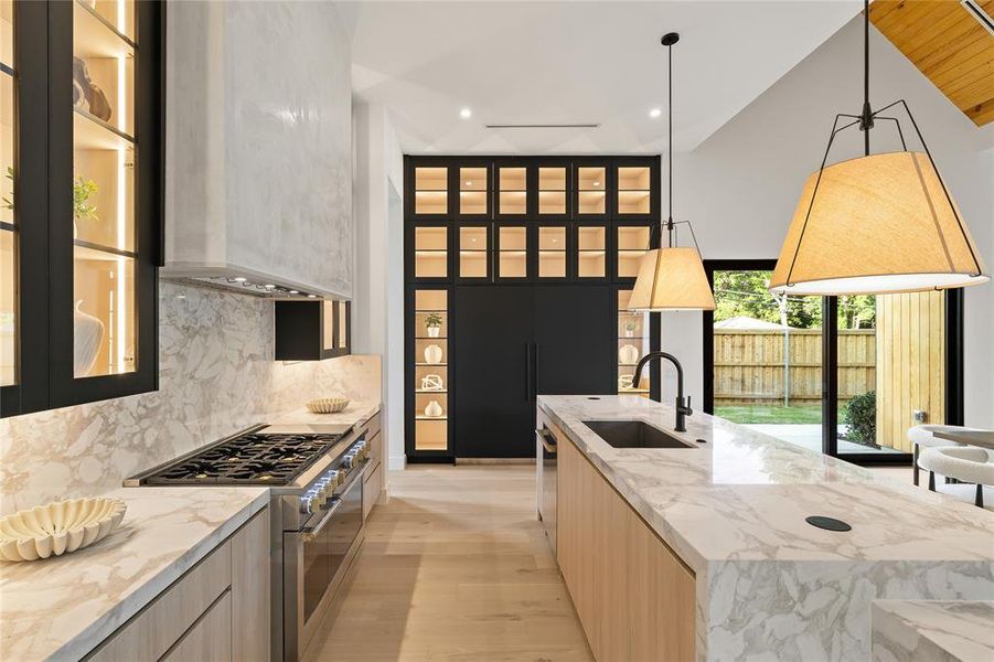 Gourmet kitchen featuring a substantial island with a built-in sink and matte black faucet, integrated stainless steel appliances, light wood-finish cabinetry, extensive marbled countertops and backsplash, and a contemporary range hood Gourmet kitchen featuring a substantial island with a built-in sink and matte black faucet, integrated stainless steel appliances, light wood-finish cabinetry, extensive marbled countertops and backsplash, and a contemporary range hood