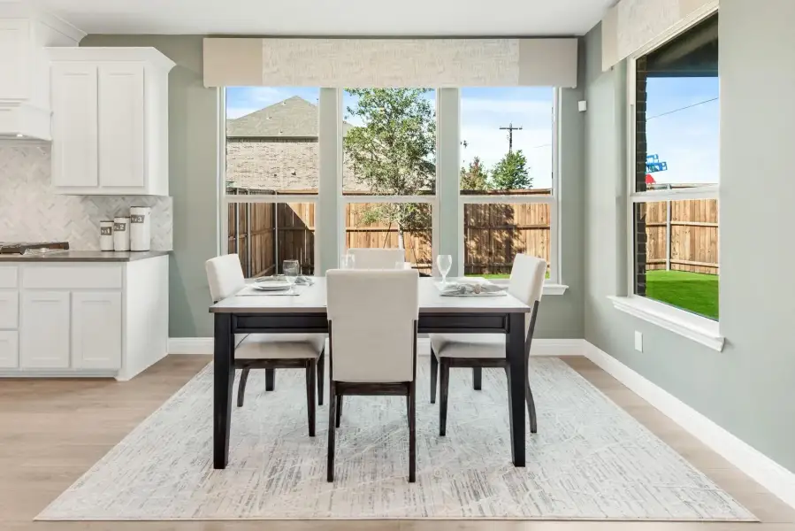 Dining area with dark wood table, four upholstered chairs, large windows, and light hardwood floors next to white kitchen cabinets