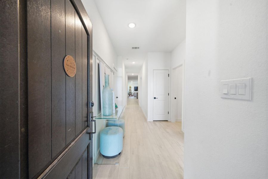 Welcome to a bright, inviting entryway featuring a wooden door and sleek flooring. The hallway is elegantly styled with modern decor and leads to spacious living areas, offering a warm and welcoming feel.
