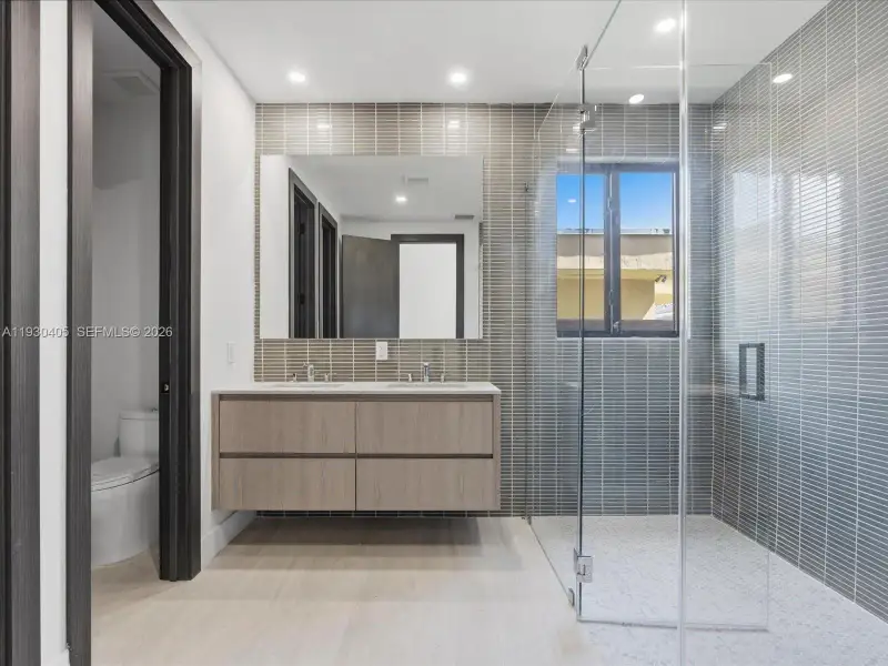 Floating dual-sink vanity with light wood-finish cabinetry, countertop with integrated dual basins, full-height tile backsplash, frameless glass-enclosed shower, and recessed ceiling lighting Floating dual-sink vanity with light wood-finish cabinetry, countertop with integrated dual basins, full-height tile backsplash, frameless glass-enclosed shower, and recessed ceiling lighting