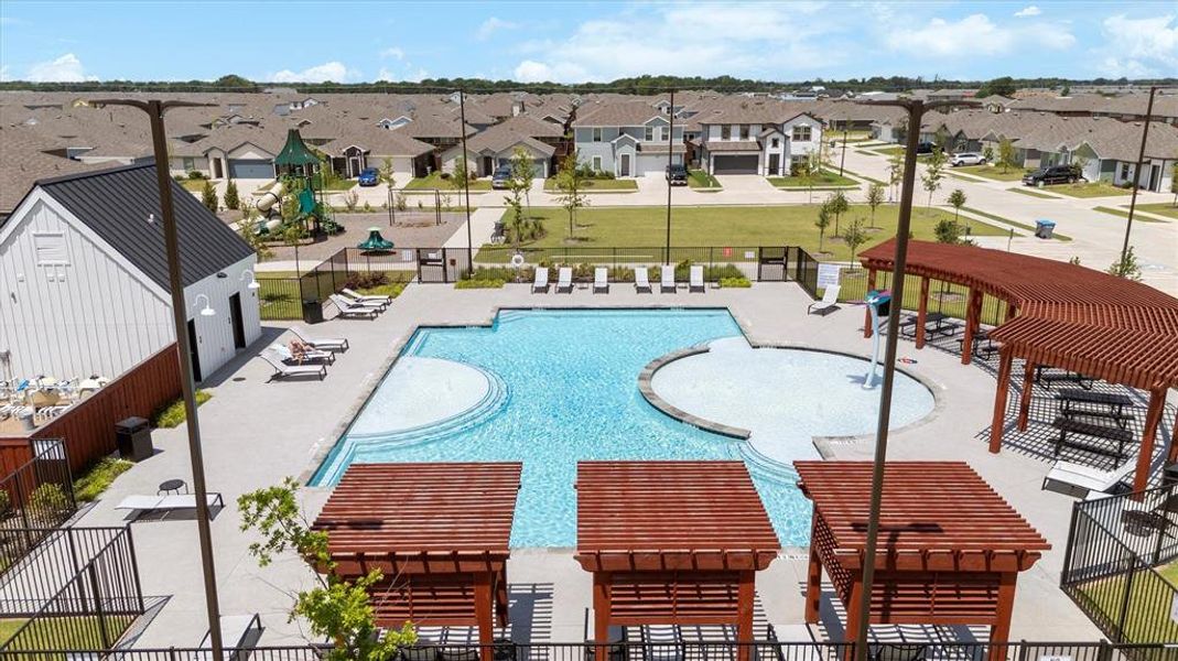 Community swimming pool with an integrated splash pad, featuring multiple sun loungers, shaded pergola structures, and a dedicated pool house with white siding and a dark metal roof