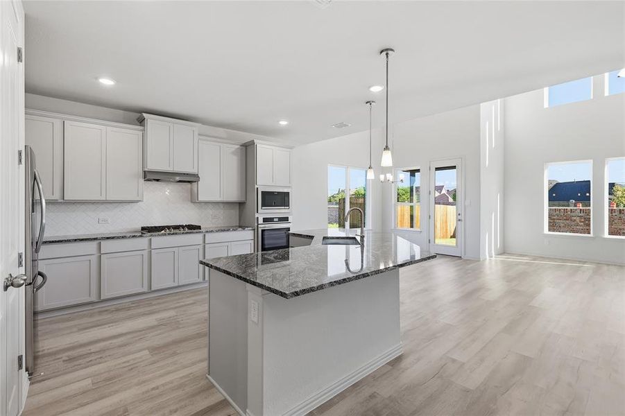 Kitchen with dark stone countertops, decorative backsplash, light wood-type flooring, an island with sink, and decorative light fixtures Kitchen with dark stone countertops, decorative backsplash, light wood-type flooring, an island with sink, and decorative light fixtures