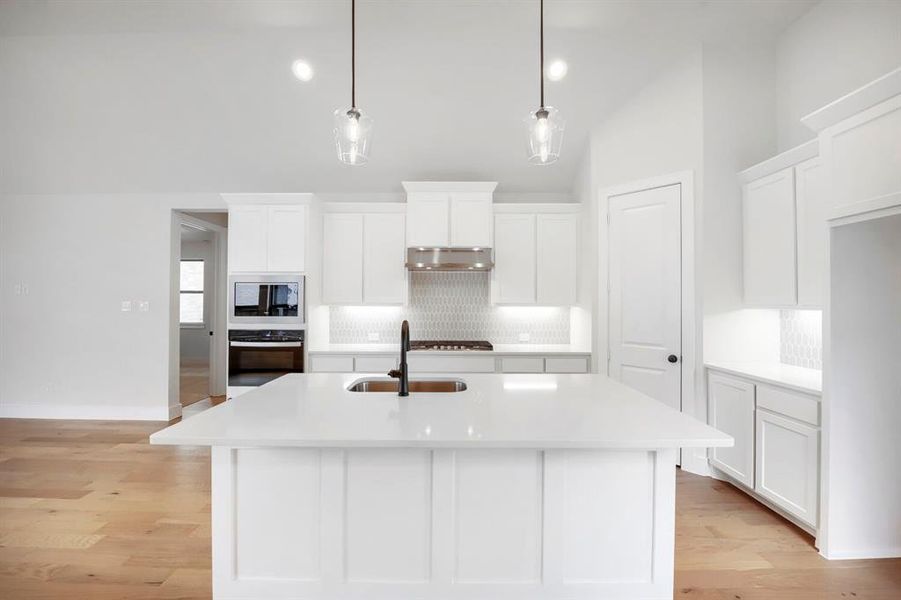 Bright kitchen featuring an expansive center island with an undermount sink, white cabinetry, integrated stainless steel appliances, and wood-finish flooring Bright kitchen featuring an expansive center island with an undermount sink, white cabinetry, integrated stainless steel appliances, and wood-finish flooring