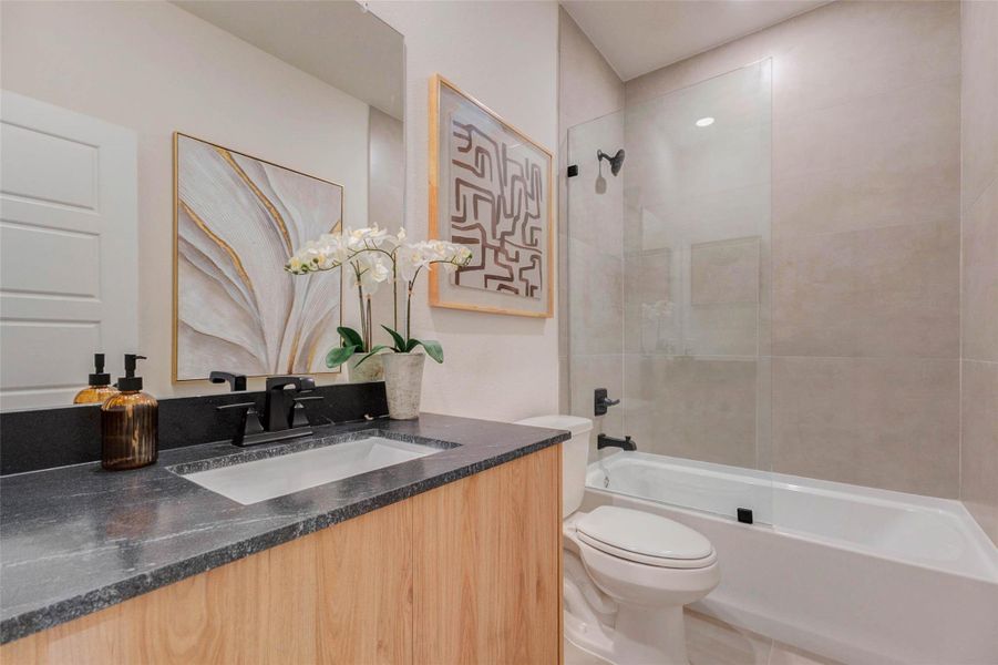 This stylish full bathroom showcases a modern blend of design and functionality, featuring a sleek vanity with dark countertops, matte black fixtures, and warm wood cabinetry for contrast. The tub and shower combination is enhanced by a glass enclosure and neutral tile surround, creating a spa-like feel. Thoughtful finishes and clean lines make this space both practical and visually appealing for everyday use. The picture shown is from the model home with the same floor plan, Rev floor plan.