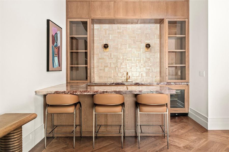 The game room features a beautifully appointed bar with custom white oak cabinetry, natural stone countertops, and a richly textured tile backsplash. Glass-front display cabinetry, integrated beverage refrigeration, and counter seating create an inviting setting for entertaining. The game room features a beautifully appointed bar with custom white oak cabinetry, natural stone countertops, and a richly textured tile backsplash. Glass-front display cabinetry, integrated beverage refrigeration, and counter seating create an inviting setting for entertaining.