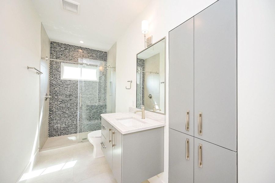 This bathroom features a modern design with sleek gray cabinetry, a large mirror, and a spacious glass-enclosed shower with a mosaic tile accent wall. Bright and well-lit, it offers a clean, contemporary feel.