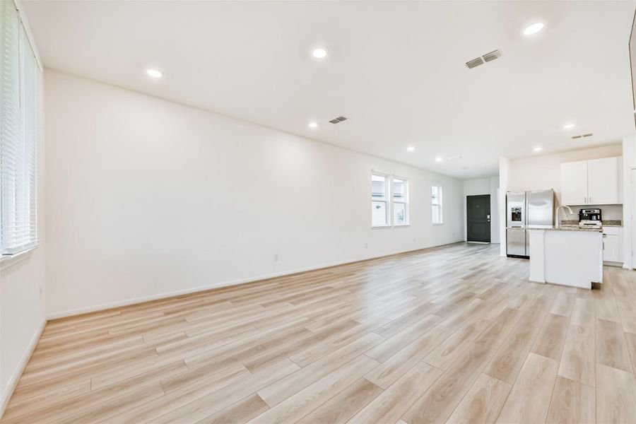 This photo showcases a spacious, open-concept living area with light wood flooring, bright walls, recessed lighting, and a modern kitchen featuring stainless steel appliances and white cabinetry. Natural light fills the room through multiple windows, creating a warm and inviting atmosphere.