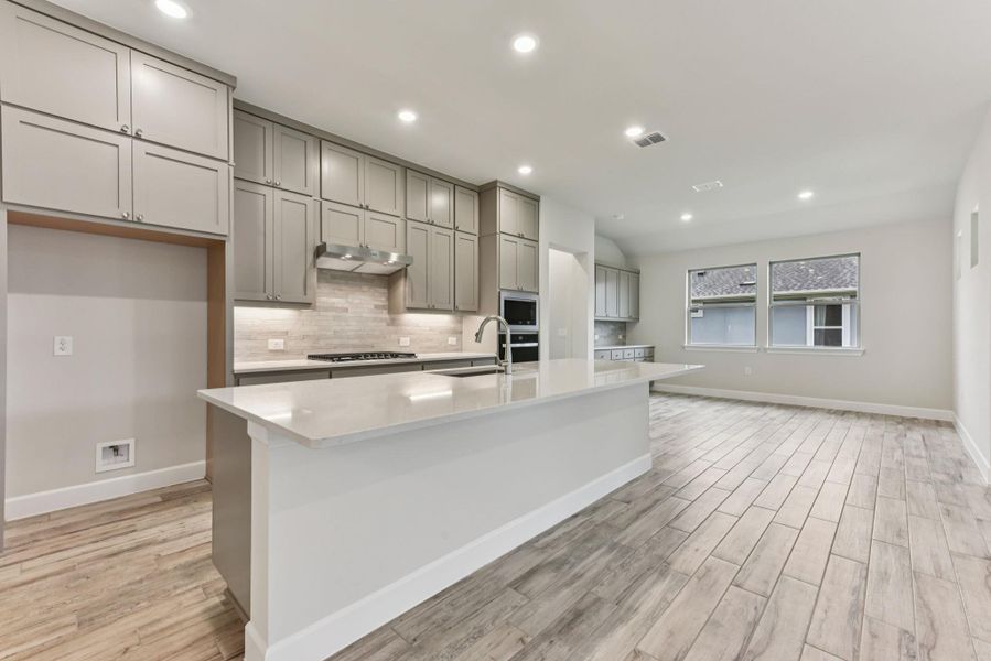 Kitchen featuring gray cabinetry, light stone counters, a center island with sink, backsplash, and recessed lighting