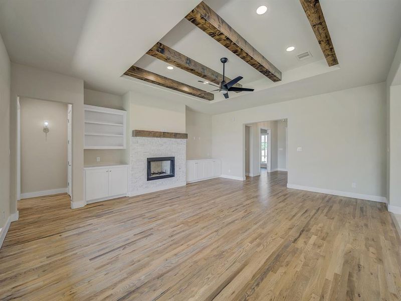 Unfurnished living room featuring ceiling fan, beam ceiling, light wood-style floors, a fireplace, and built in features Unfurnished living room featuring ceiling fan, beam ceiling, light wood-style floors, a fireplace, and built in features