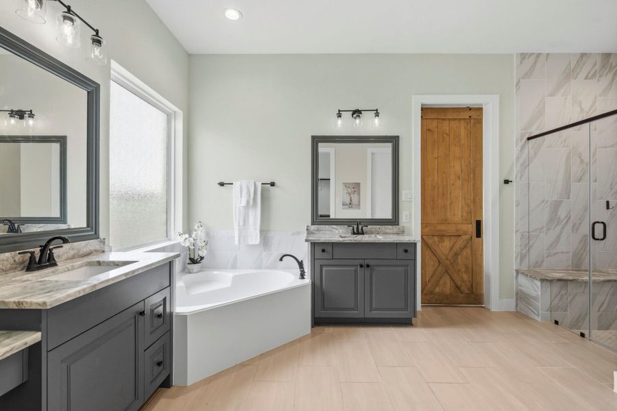A relaxing corner garden tub serves as a focal point, positioned beneath a large translucent window that fills the space with natural light while maintaining privacy. The oversized frameless shower features a floor-to-ceiling marble-inspired tile and a granite-topped bench seat. Completing this exceptional retreat are two oversized walk-in closets, providing abundant storage and organization while enhancing the suite’s sense of elegance and functionality.