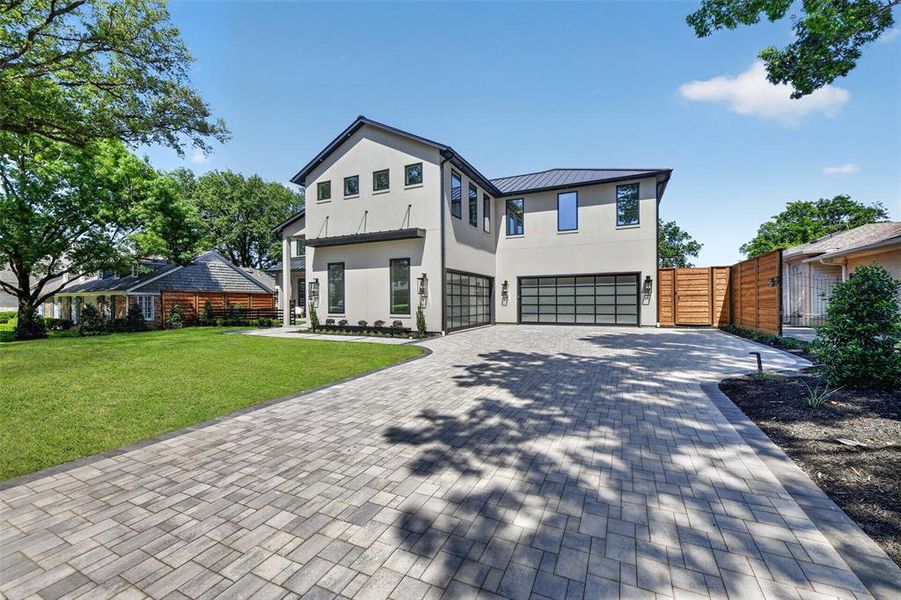 Contemporary stucco exterior with a dark metal roof, featuring a paver driveway, two-car garage with frosted glass doors, a manicured front lawn, and a wood privacy fence Contemporary stucco exterior with a dark metal roof, featuring a paver driveway, two-car garage with frosted glass doors, a manicured front lawn, and a wood privacy fence