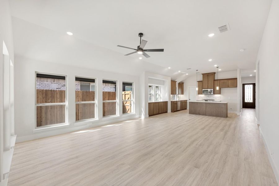 Open living area with light wood floors, ceiling fan, and kitchen with brown cabinets and island in background