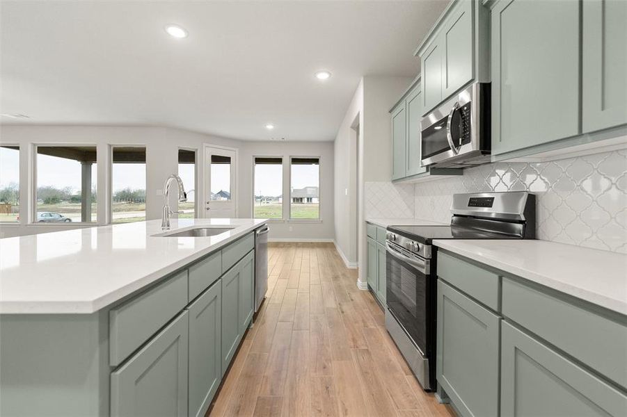 Kitchen featuring stainless steel appliances, a kitchen island with sink, tasteful backsplash, light stone countertops, and light wood-type flooring Kitchen featuring stainless steel appliances, a kitchen island with sink, tasteful backsplash, light stone countertops, and light wood-type flooring
