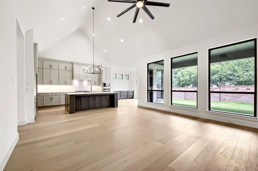 Open-concept living space featuring high vaulted ceilings, light wood-finish flooring, a contemporary kitchen island with seating, pendant lighting, and multiple large windows overlooking exterior landscaping Open-concept living space featuring high vaulted ceilings, light wood-finish flooring, a contemporary kitchen island with seating, pendant lighting, and multiple large windows overlooking exterior landscaping