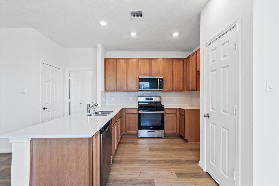 The kitchen boasts high ceilings, recessed lighting, Woodmont Cody cabinets in a Toffee stain, Silestone Blanco Maple countertops, stainless steel Whirlpool appliances, a modern Daltile backsplash, and a pantry—all overlooking the Great Room.