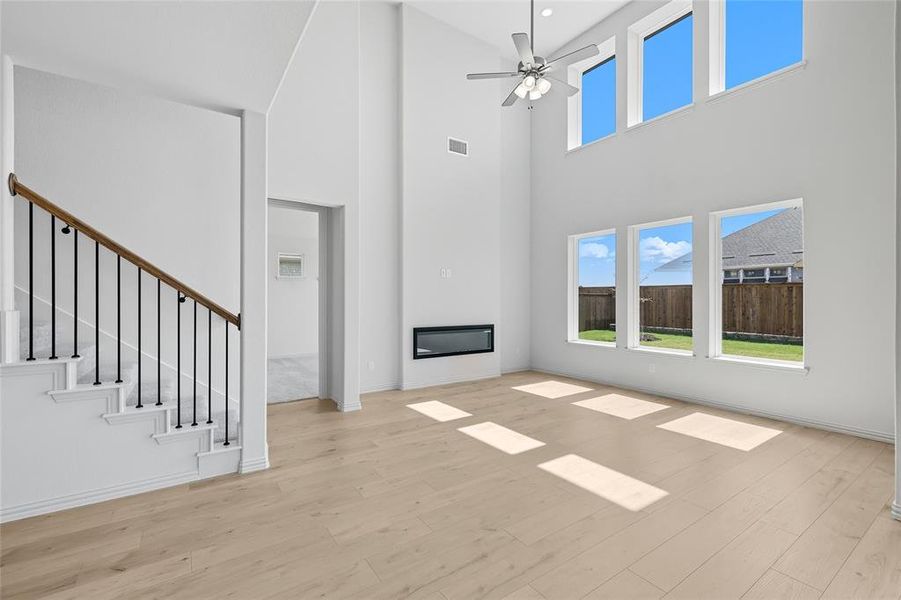 Unfurnished living room with a towering ceiling, a glass covered fireplace, ceiling fan, light wood finished floors, and stairs Unfurnished living room with a towering ceiling, a glass covered fireplace, ceiling fan, light wood finished floors, and stairs
