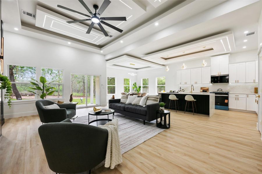 Open-concept living space featuring high ceilings with integrated lighting and decorative tray details, wood-finish flooring, a contemporary ceiling fan, and large windows with a sliding glass door providing abundant natural light