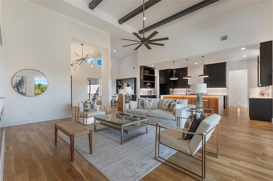 Living room with ceiling fan with notable chandelier, a high ceiling, beam ceiling, and light wood-type flooring