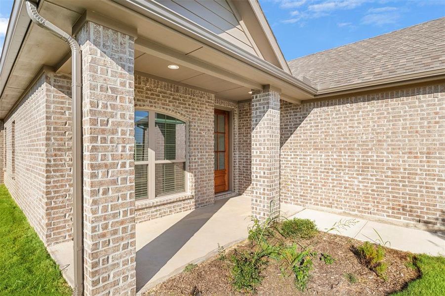 View of exterior entry featuring brick siding, roof with shingles, and a patio area