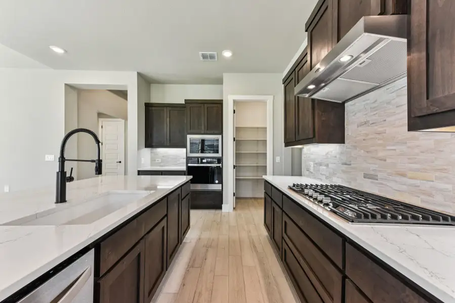 Kitchen with dark brown cabinetry, backsplash, light stone countertops, exhaust hood, and stainless steel appliances